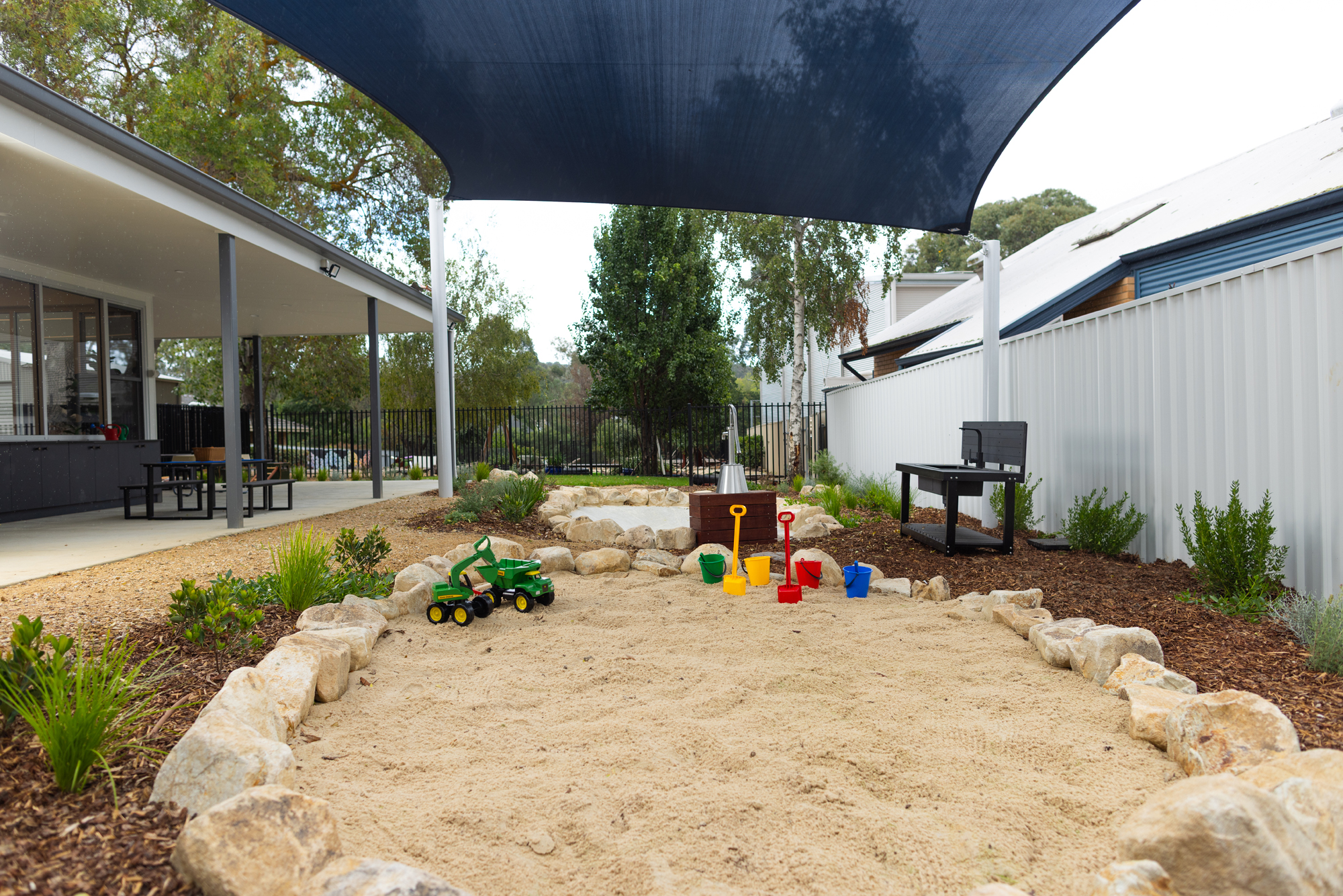 St Michael’s Early Learning Centre outdoor sand play area with a shaded play space, rocks, toys, and natural landscaping