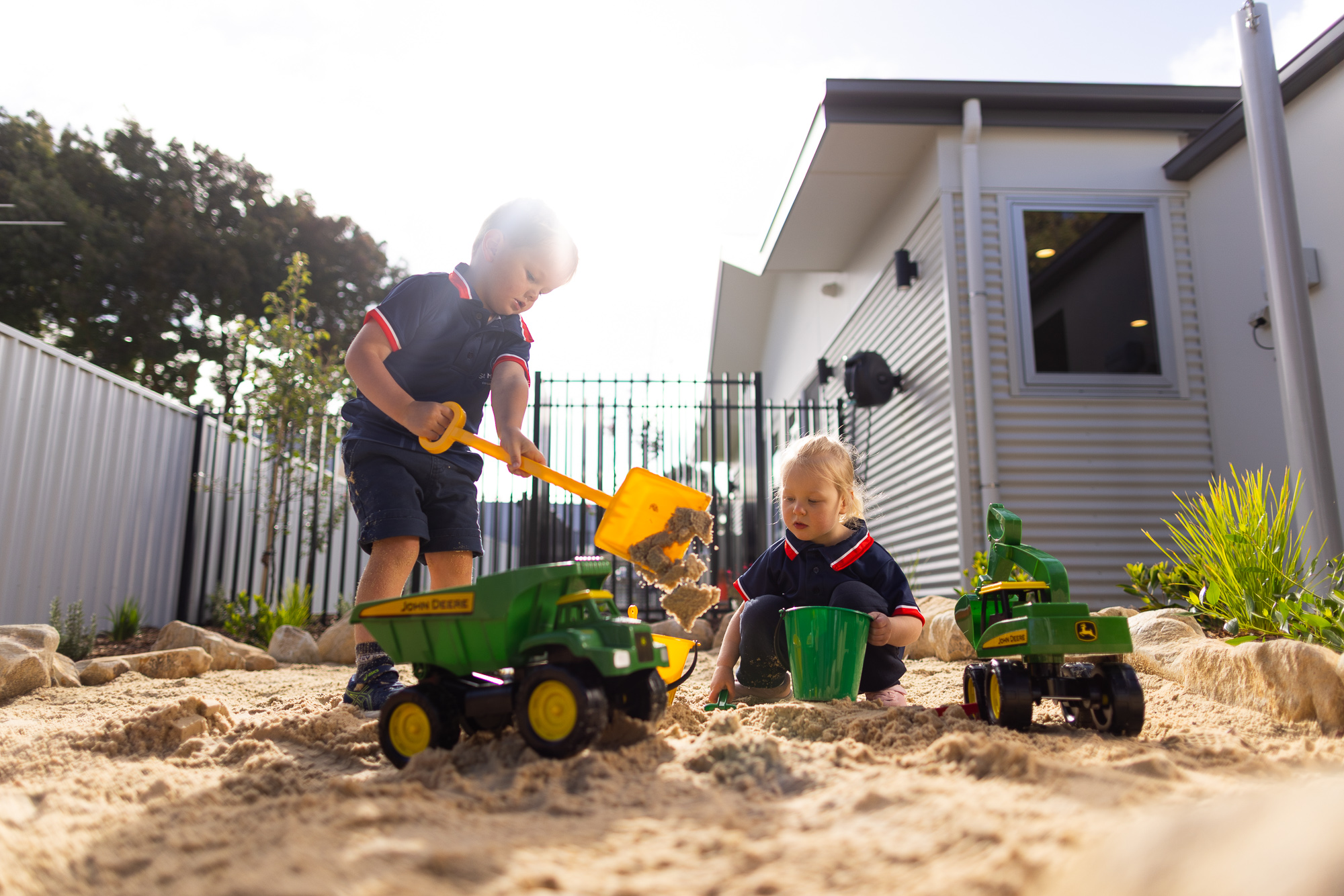 Children playing with sand and toy trucks in the outdoor play area at St Michael’s Early Learning Centre. > Young students playing with toy trucks and digging in a sandpit at an outdoor early learning area.