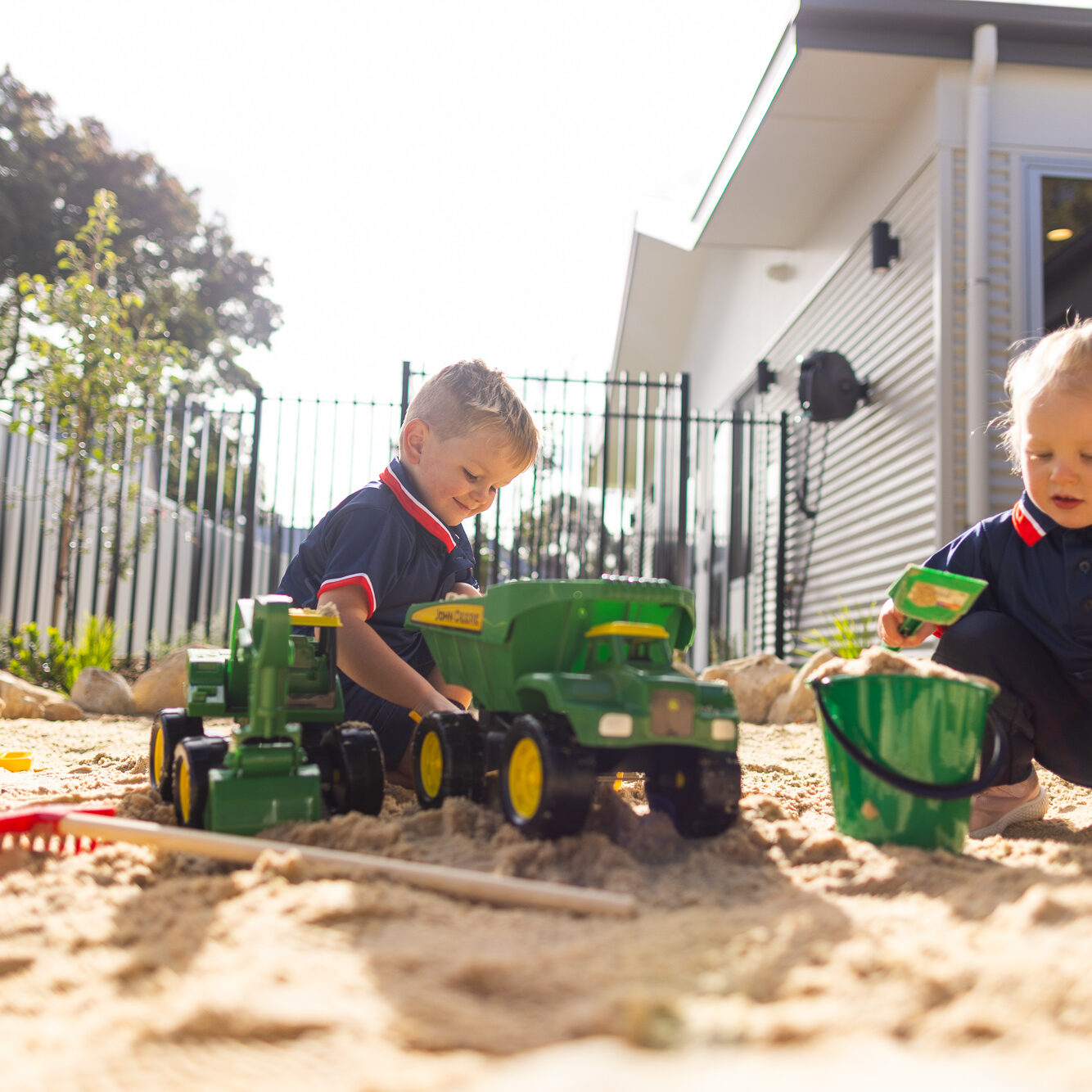 Two young children in St Michael’s Early Learning Centre uniforms playing with toy trucks and a bucket in an outdoor sandpit.
